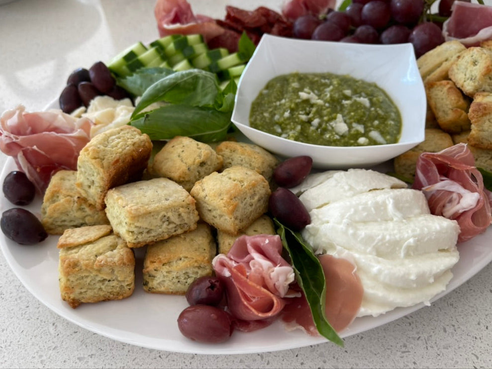 Platter with assorted finger foods including Pesto Biscuitini, cured meats, and olives on a white surface.