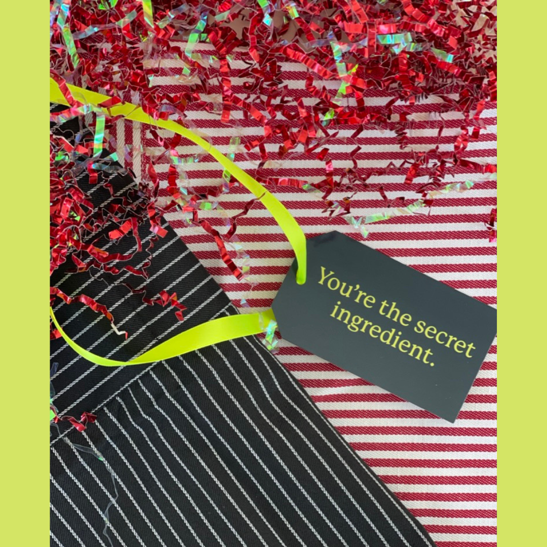 Gift with black and white striped aprons, red crinkle paper, and a tag saying 'You're the secret ingredient' on red and white striped aprons.