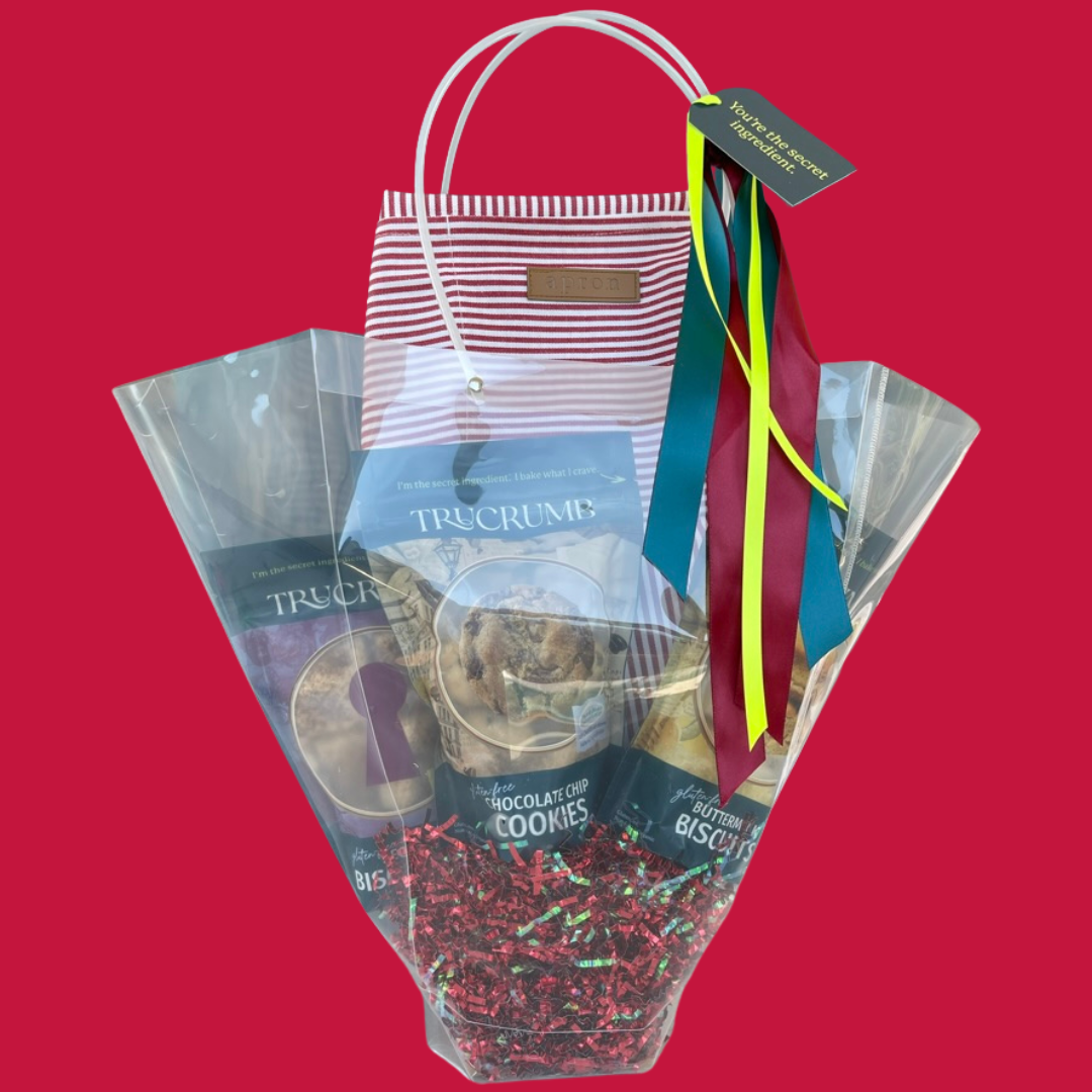 Gift bag with TruCrumb gluten free cookies, biscuits, apron and charm and a colorful ribbon on a red background