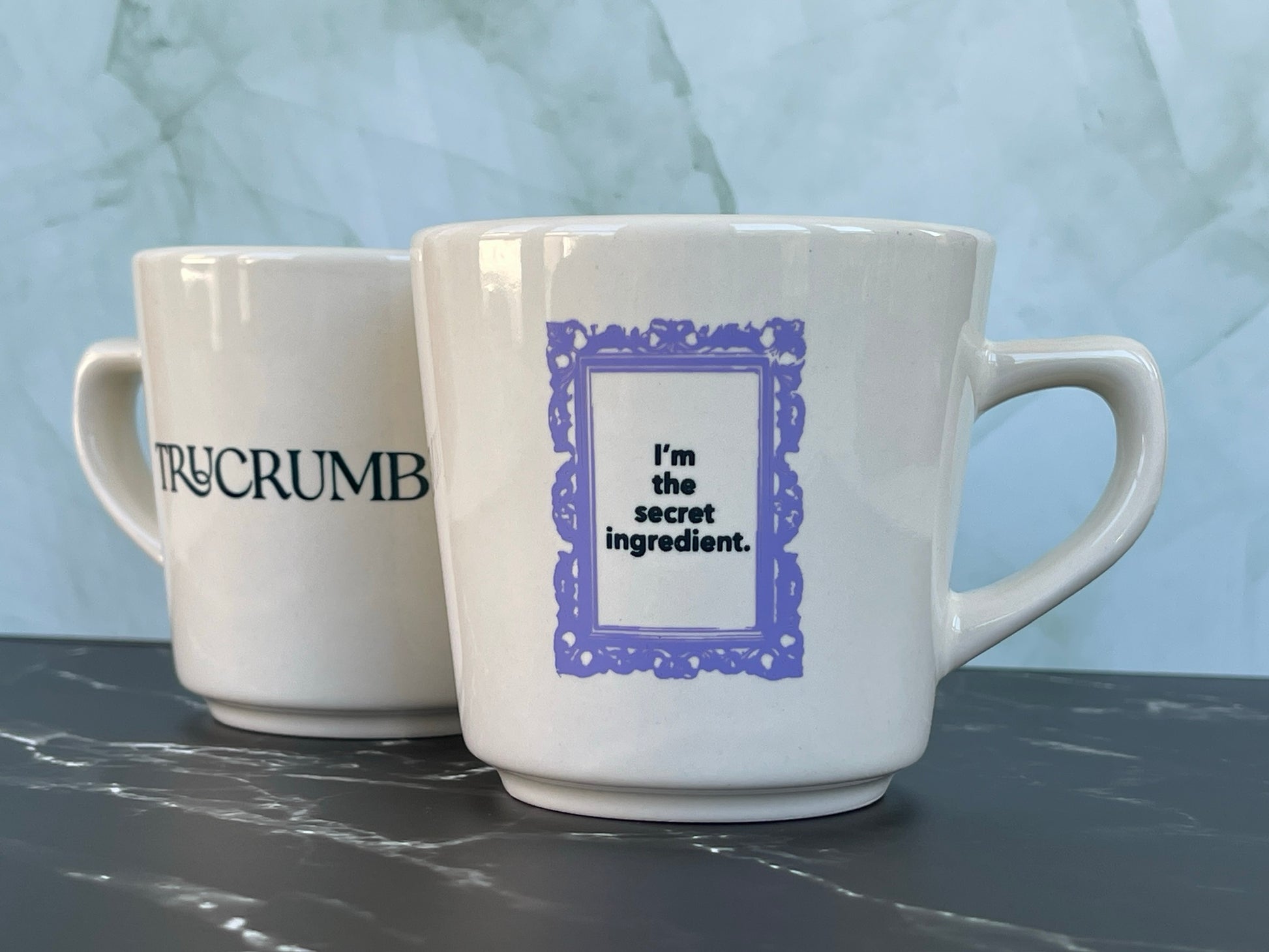 Two TruCrumb white mugs with text on a marble surface